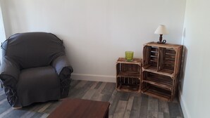 A room with a grey armchair, a wooden side table with a lamp, and a small stool. - Maison au Calme Avec Jardin, Terrasse et Barbecue (Dampierre-sur-Boutonne)