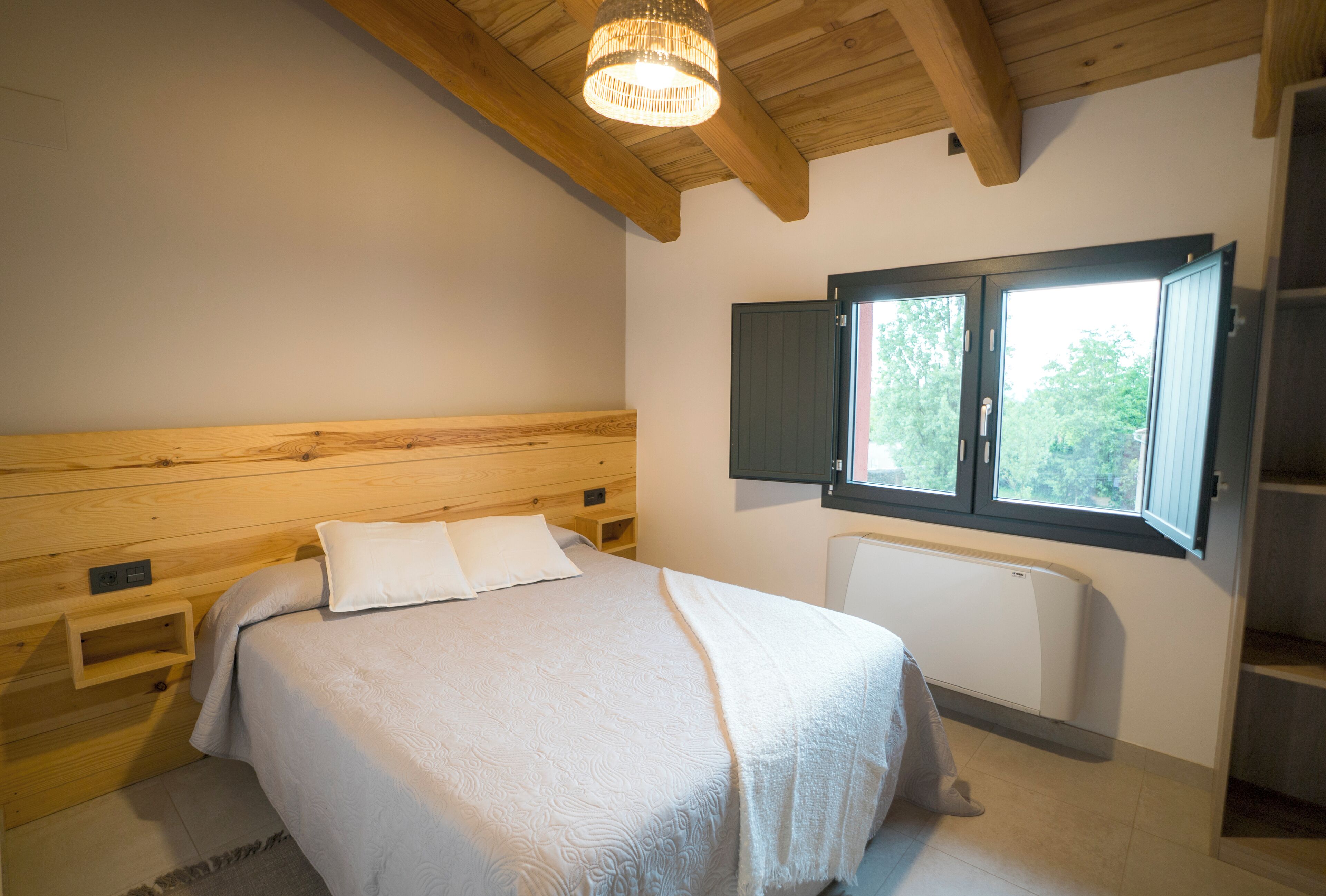 A bedroom with a wooden headboard, a bed with white bedding, a window with a view of greenery, and a built-in heater.