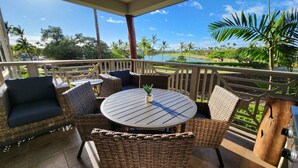 A covered outdoor seating area with wicker furniture and a round table, overlooking a scenic landscape with palm trees and a body of water.