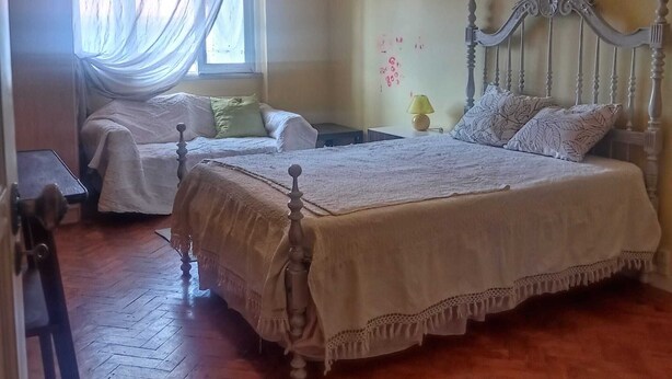 A bedroom with two beds, a window with sheer curtains, a bedside table with a lamp, and a wooden floor. - Room for Rent in Almada/lisbon (Almada)