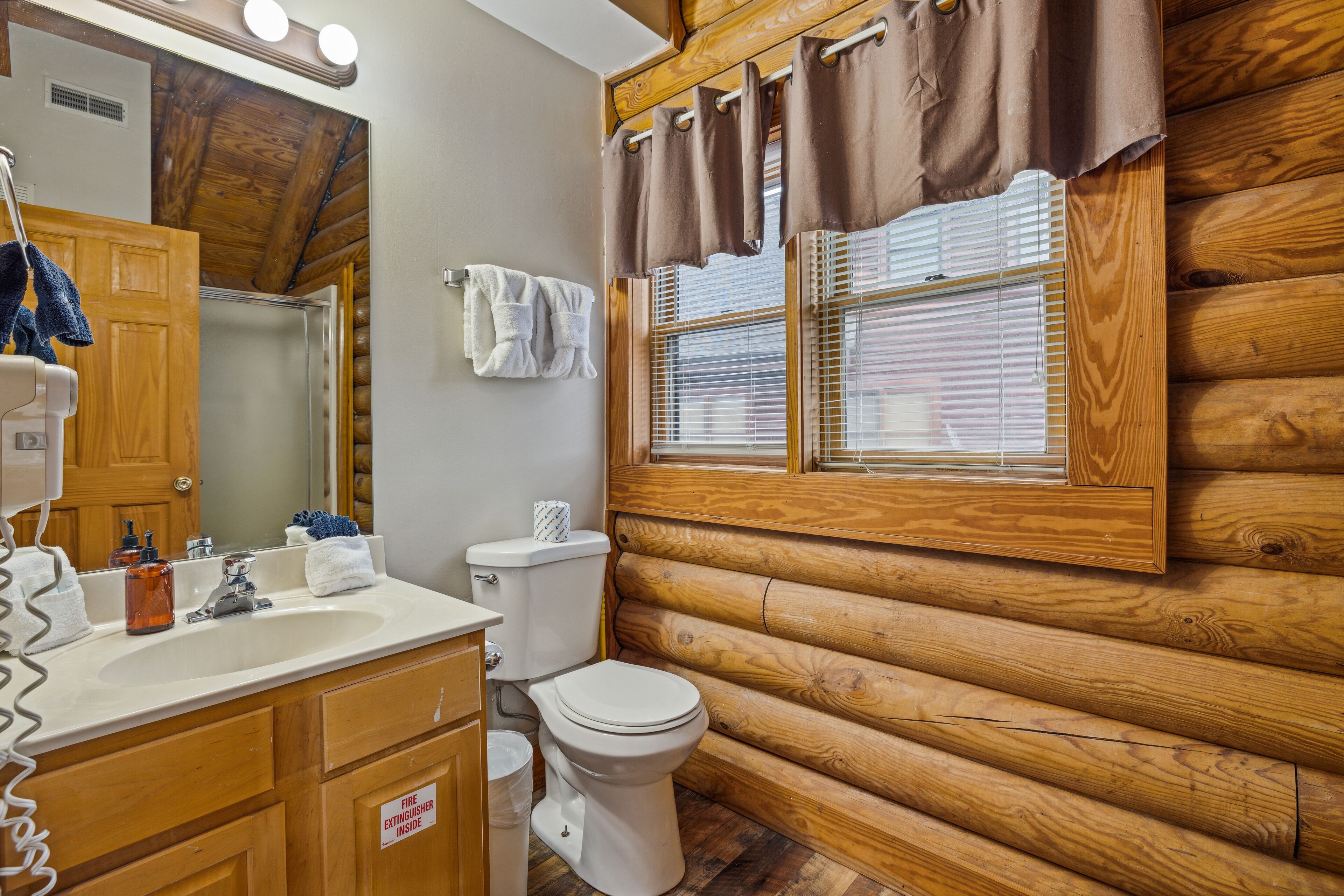 A bathroom with a wooden log cabin interior, a sink, toilet, and a window with blinds.