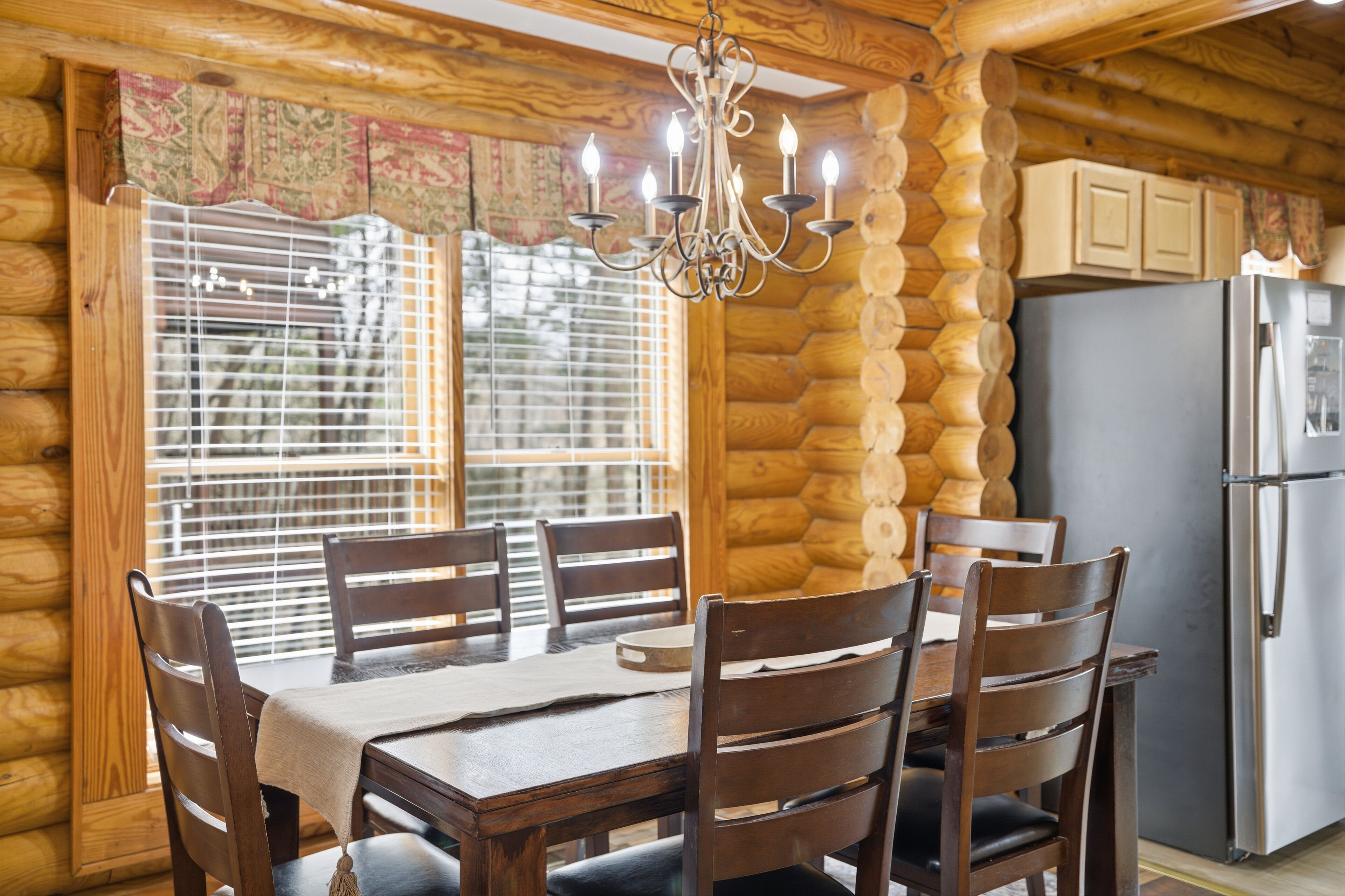 A wooden cabin interior with a dining table, chairs, a chandelier, and a refrigerator.