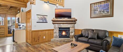 A cozy living room with a stone fireplace, a wooden ceiling, and a black leather sofa.
