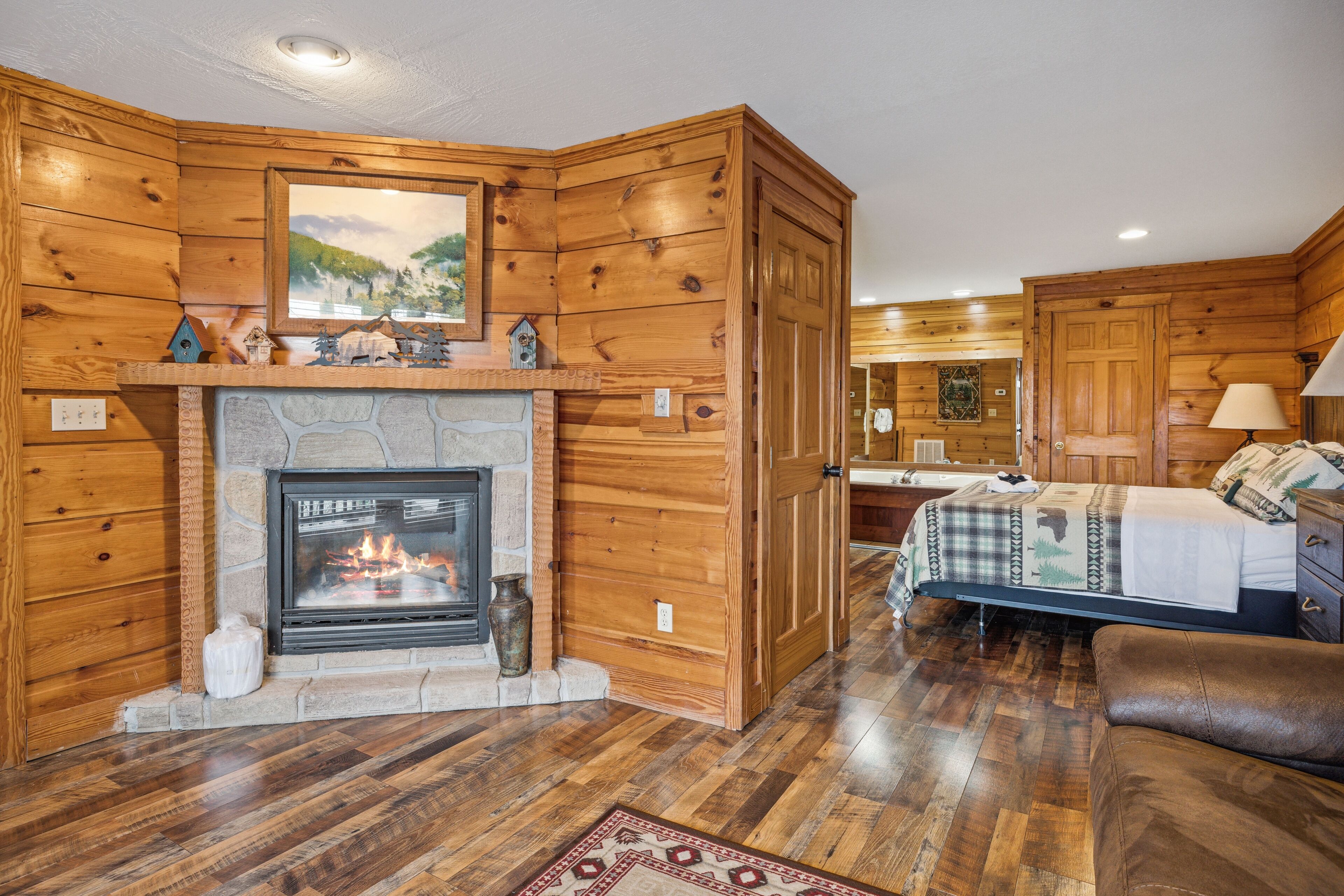 A log cabin interior with a stone fireplace, wooden walls, a bed with a patterned bedspread, and a brown leather couch.