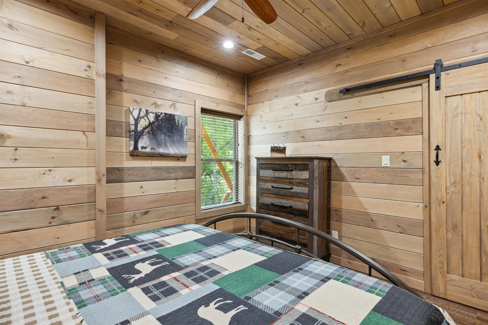 A wooden cabin room with a bed, a window, a painting, and a chest of drawers.
