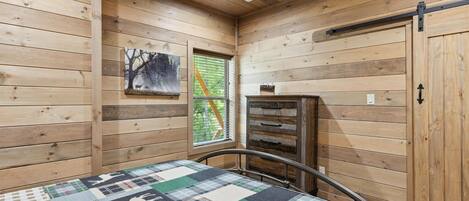 A wooden cabin room with a bed, a window, a painting, and a chest of drawers.