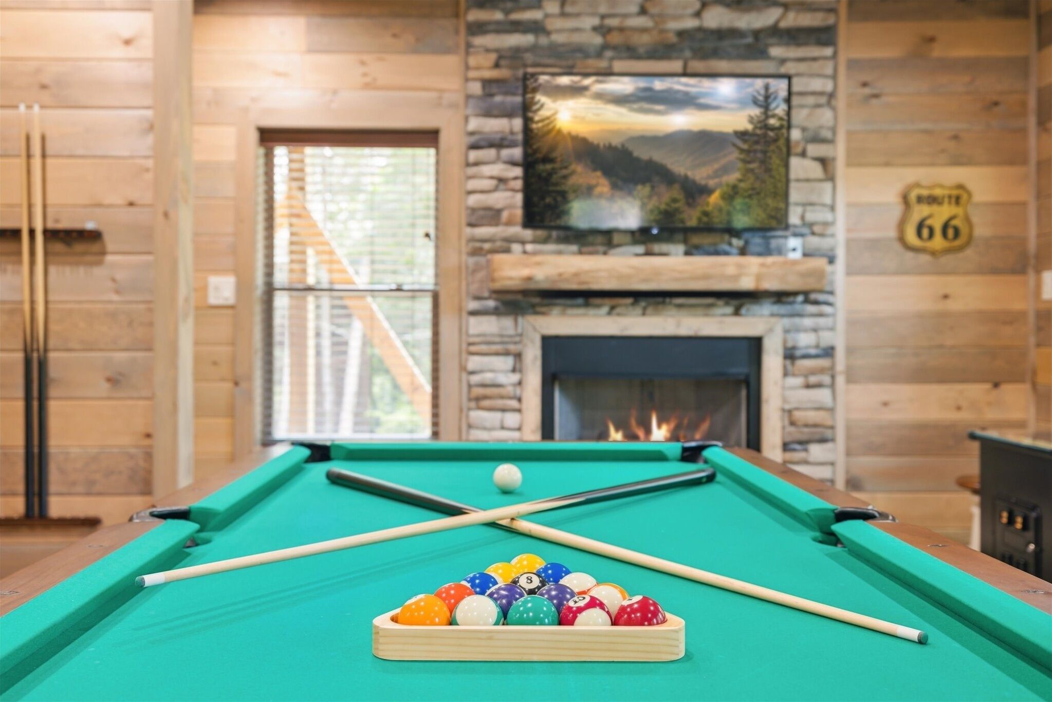 Une table de billard recouverte d’un drap vert, un jeu de queues de billard et un porte-billes, le tout dans une pièce aux murs en bois et dotée d’une cheminée en pierre.