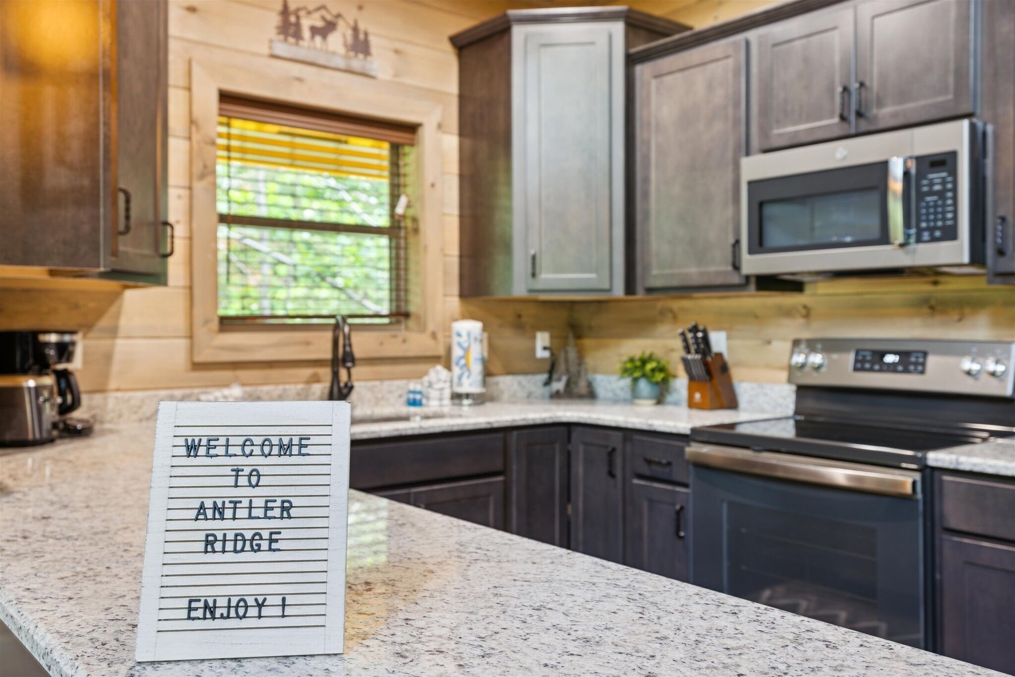 A kitchen with dark cabinets, a microwave, and a stove. A welcome sign is on the counter.