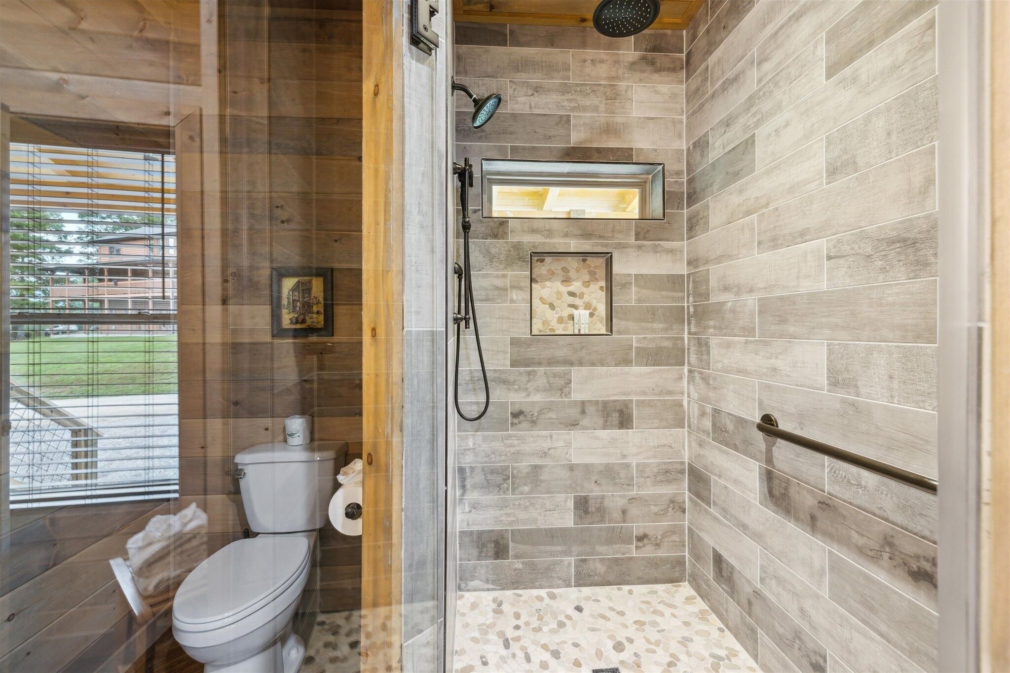 A bathroom with a wooden floor, a toilet, a shower with a glass door, and a window with blinds.