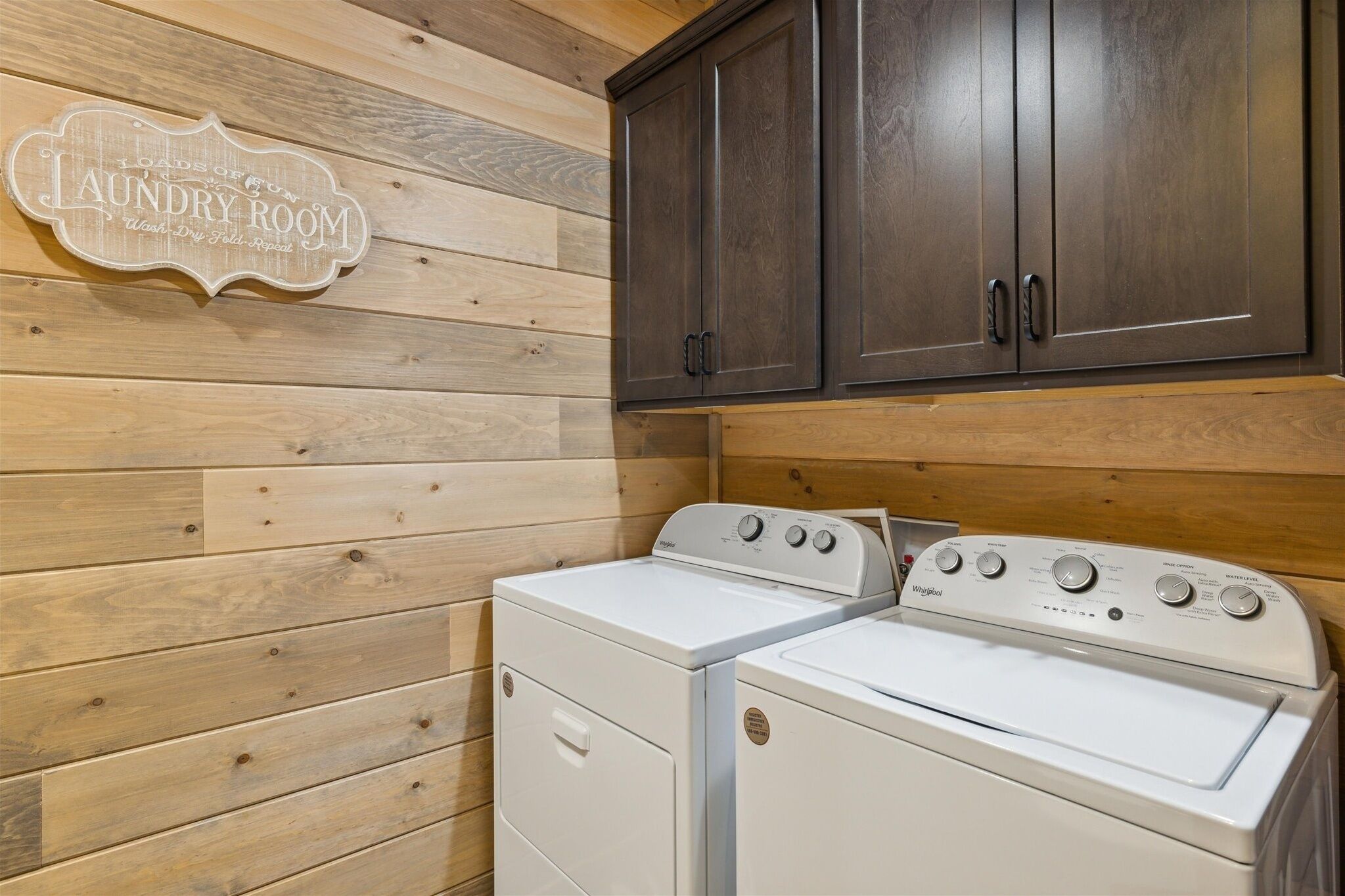 A laundry room with wooden panel walls, a white washer and dryer, and dark wooden cabinets above.