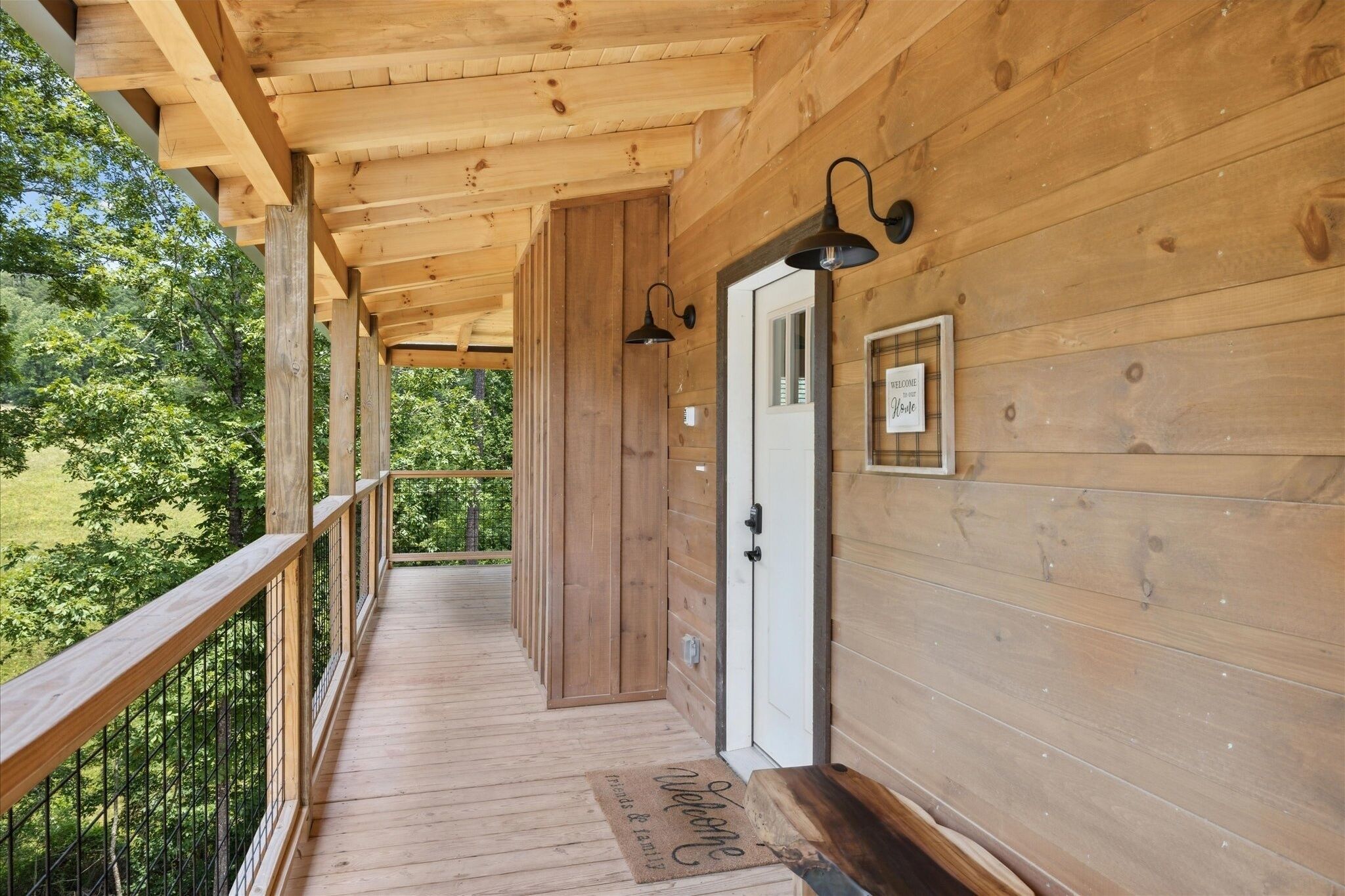 A wooden porch with a wooden railing, a door with a welcome sign, and a view of trees.
