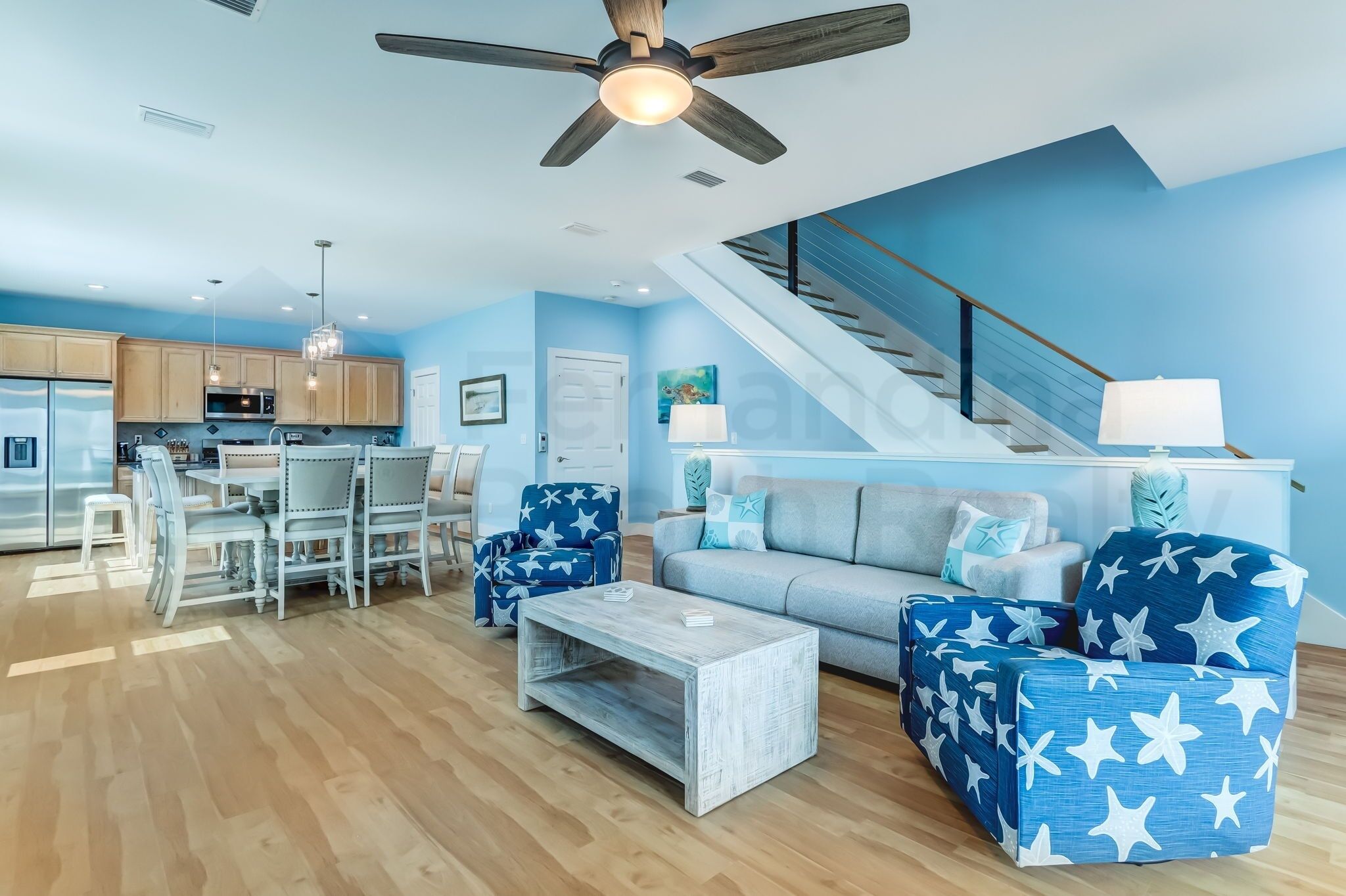 A modern living room with a sofa, coffee table, and chairs, a kitchen with wooden cabinets, and a staircase.