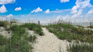 A sandy path leading through a grassy area with a clear blue sky and clouds.