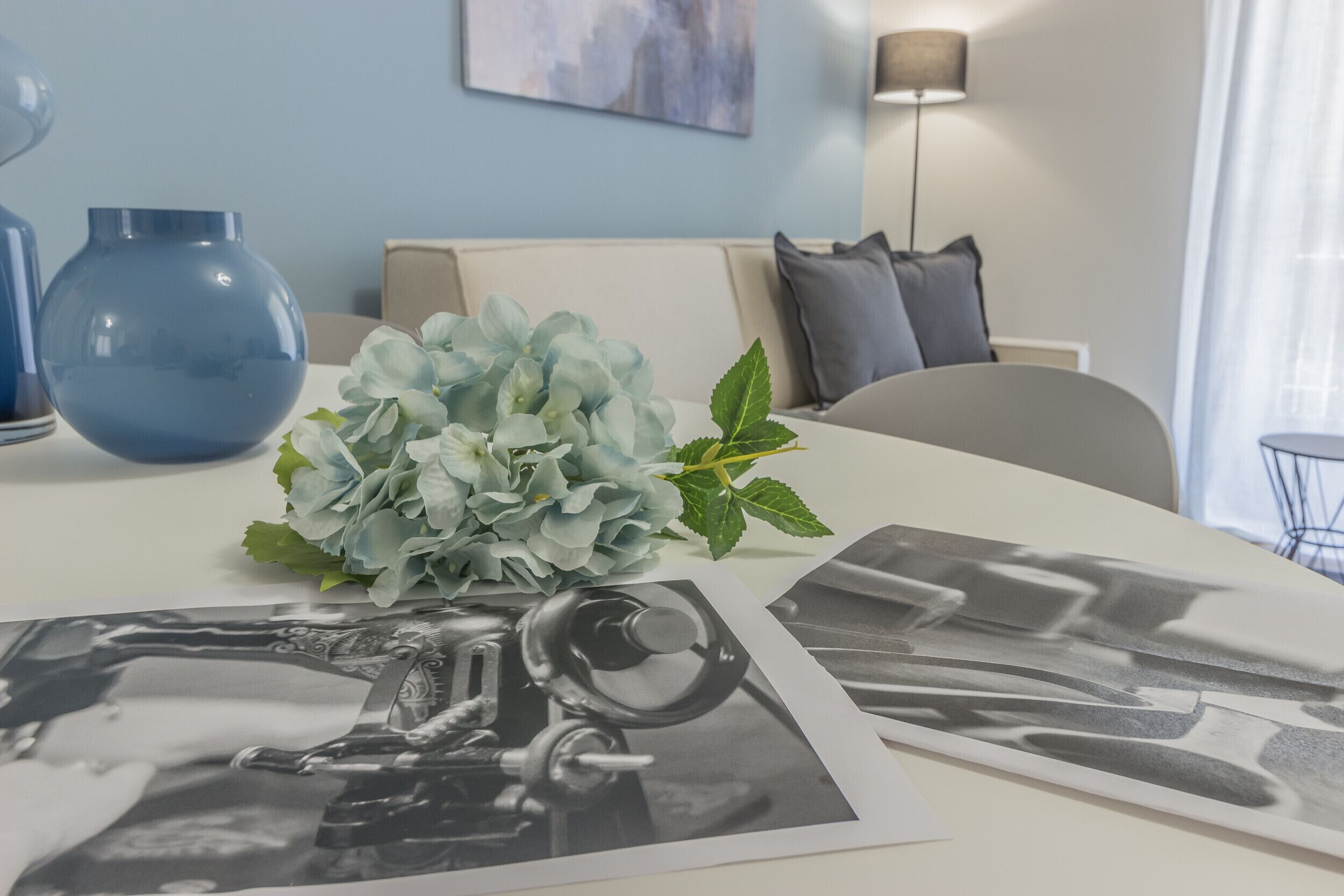 Una mesa blanca con un florero azul, un ramo de flores azul claro y fotografías en blanco y negro.