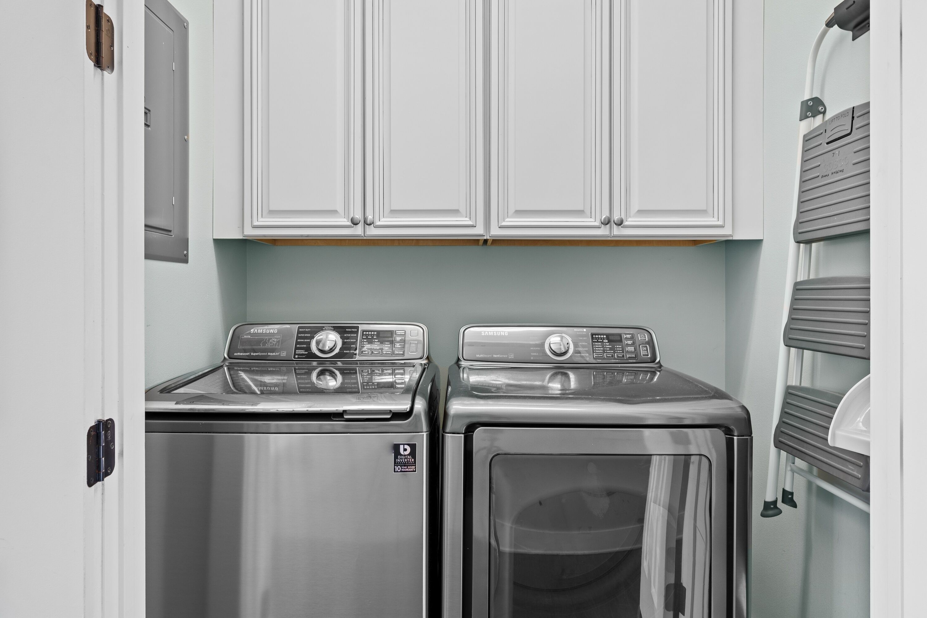A compact laundry room with two stainless steel front-loading washers and dryers, white cabinetry above, and a gray utility wall on the right.