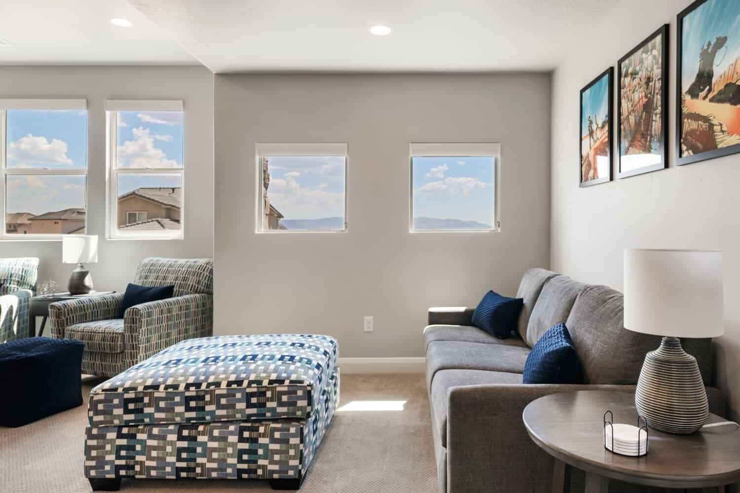 A modern living room with a sofa, armchair, ottoman, and a table with a lamp. There are three windows with views of the outside.