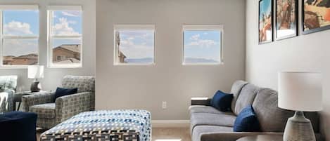 A modern living room with a sofa, armchair, ottoman, and a table with a lamp. There are three windows with views of the outside.