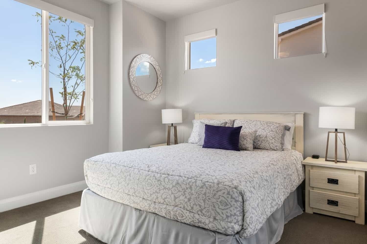 A bedroom with a large bed, two bedside tables, a nightstand, a mirror, and a window with a view of a tree and buildings.