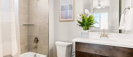 Une salle de bain moderne avec une baignoire, des toilettes et une douche. On y trouve un lavabo avec un miroir au-dessus, et une plante posée sur le comptoir.
