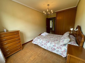 A bedroom with a wooden dresser, a bed with floral bedding, a nightstand with a lamp, and a wooden wardrobe.