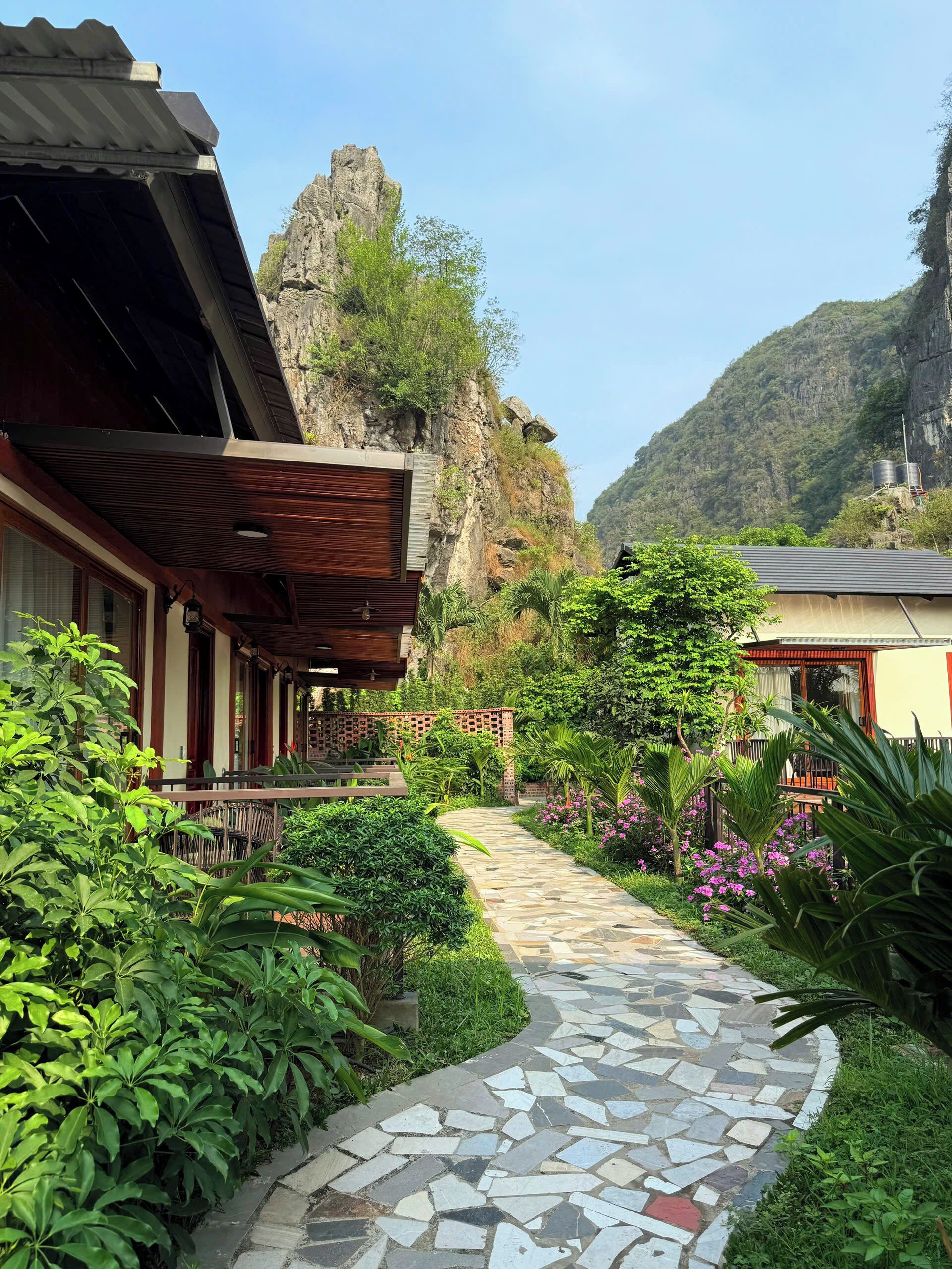 A pathway leading to a building with a wooden roof and a brick wall, surrounded by lush greenery and a mountainous backdrop.