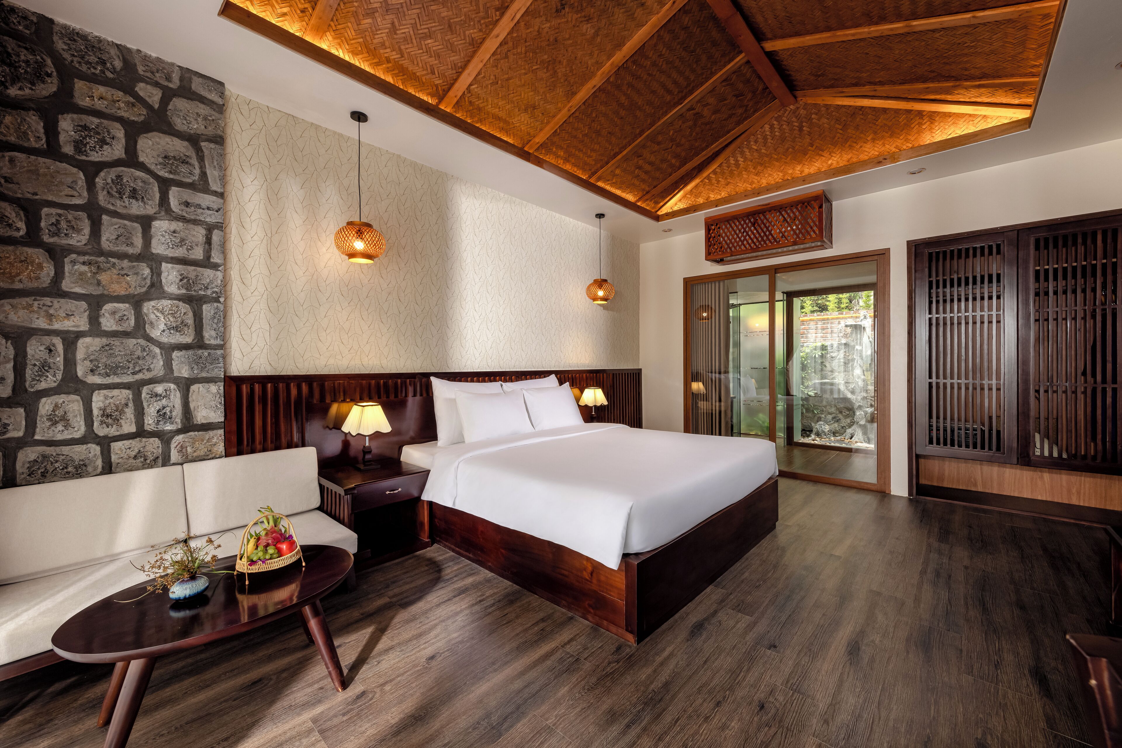 A modern hotel room with a large bed, wooden headboard, stone wall, and a view of the outdoors.