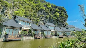 A row of modern villas with balconies overlooking a calm body of water, set against a backdrop of lush green cliffs. - Athena Premier Resort Ninh Bình (Nam Hoa Lu)