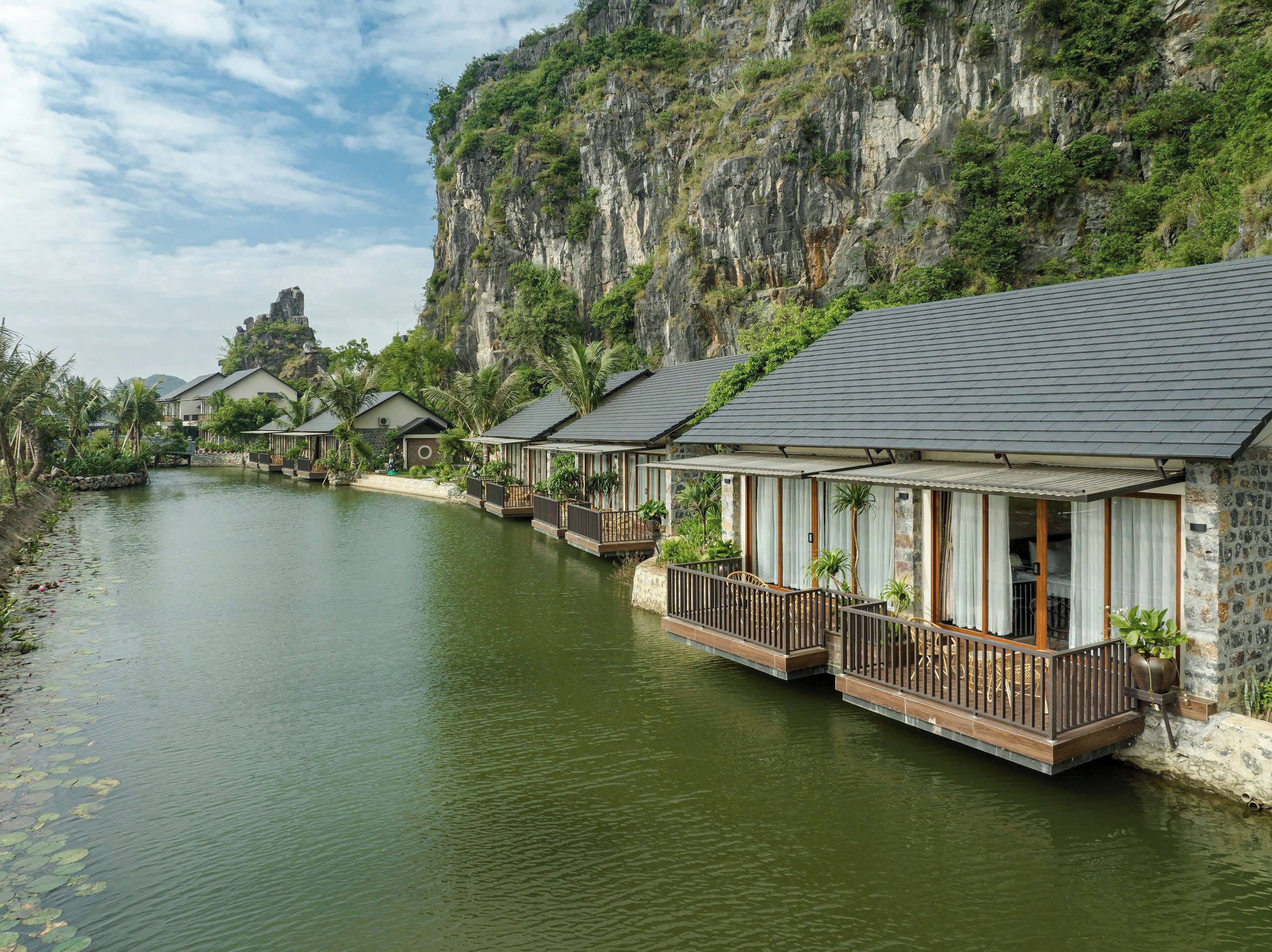 A row of bungalow-style accommodations with balconies overlooking a calm river, set against a backdrop of a steep cliff.