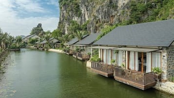 A row of bungalow-style accommodations with balconies overlooking a calm river, set against a backdrop of a steep cliff.
