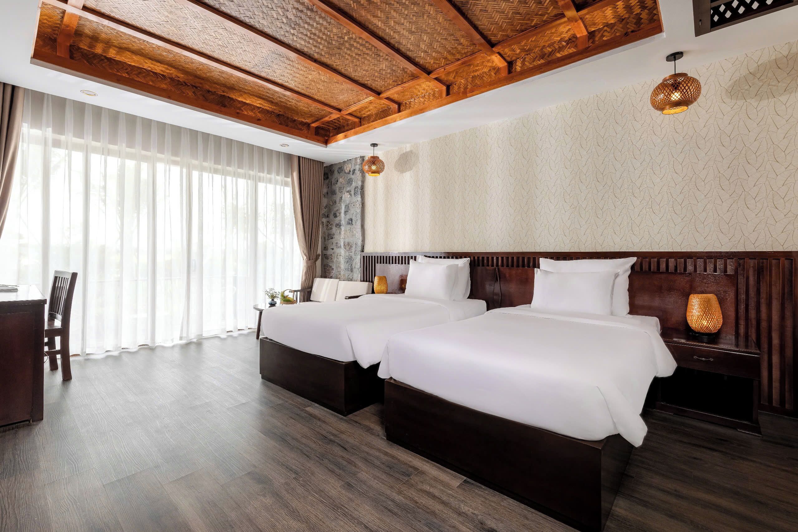 Habitación de hotel con dos camas, techo de madera, un ventanal grande con cortinas y una mesita con una silla.