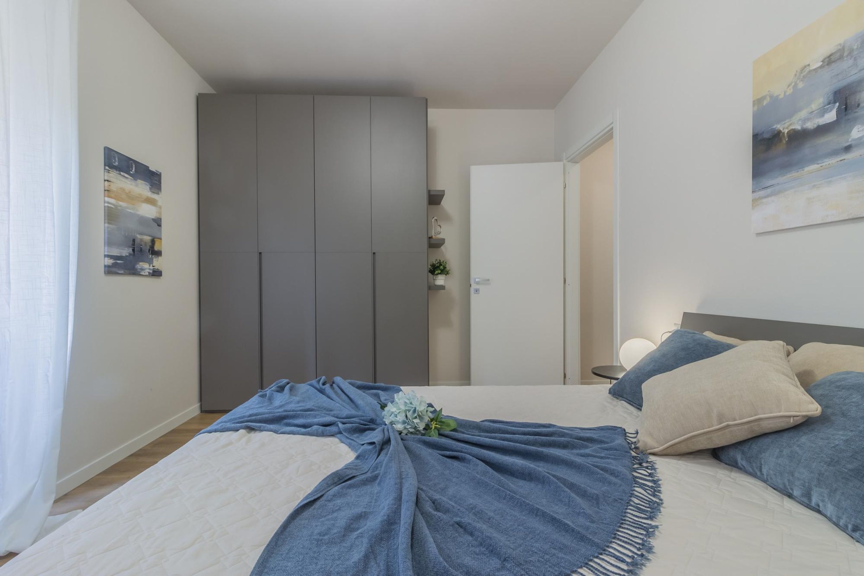 Une chambre à coucher avec un lit, une armoire grise, une porte et un mur orné d’un tableau.