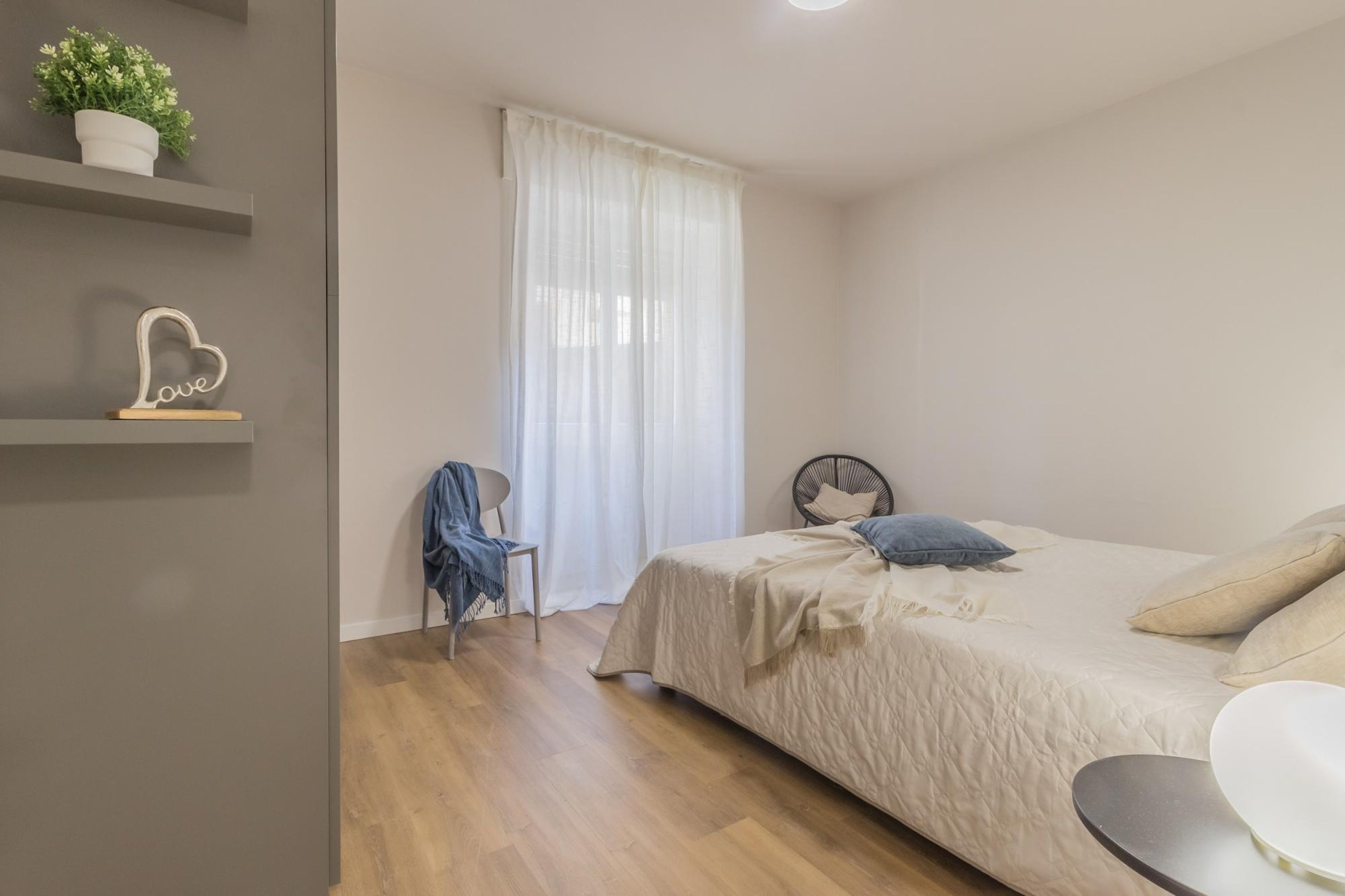 A bedroom with a bed, a chair, a shelf with a plant, and a window with curtains.