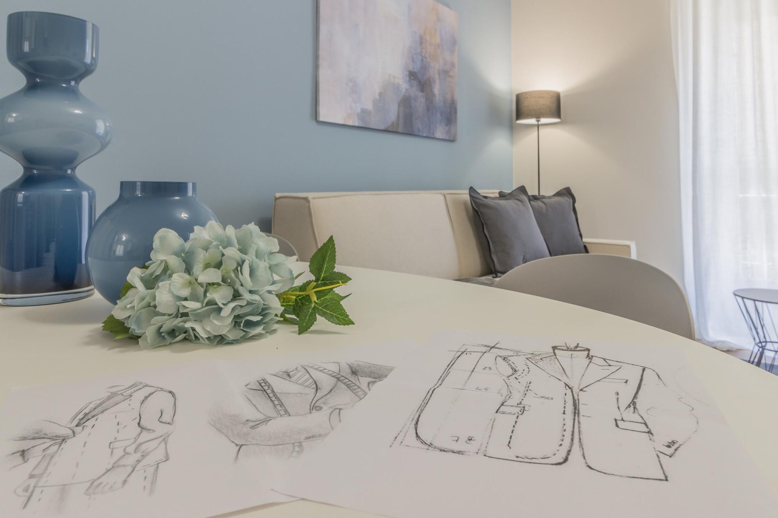 Una habitación con una mesa blanca que presenta bocetos de diseños de ropa, un jarrón con flores y dos jarrones azules.