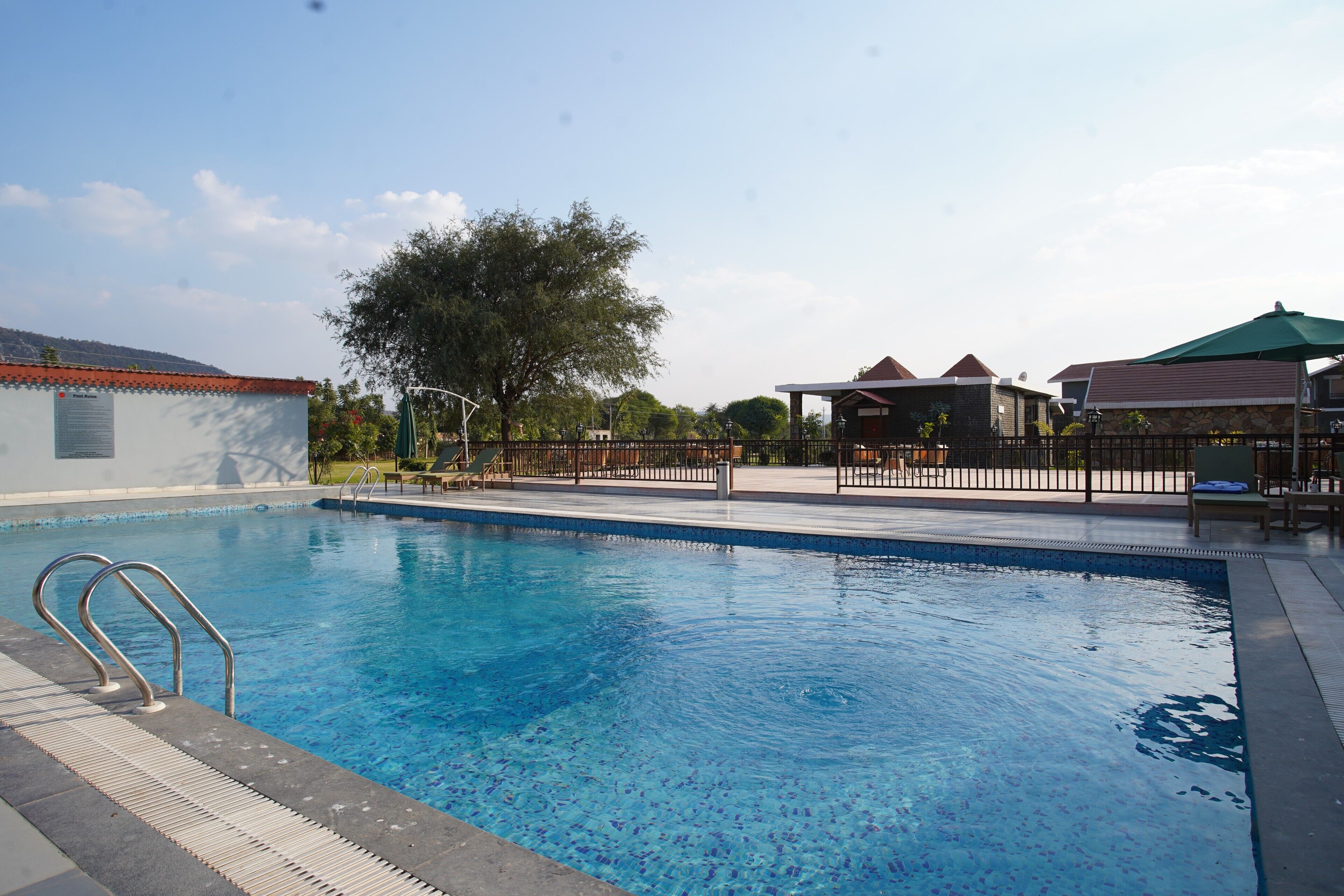 A swimming pool with a metal ladder, a covered seating area, and a building with a red roof in the background.