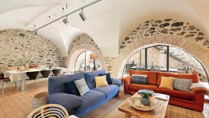 A cozy living area with a blue sofa, an orange sofa, and a wooden coffee table. There is a dining table with chairs and a stone wall in the background. - Garriguella- Roses – Pool Costa Brava House (Garriguella)