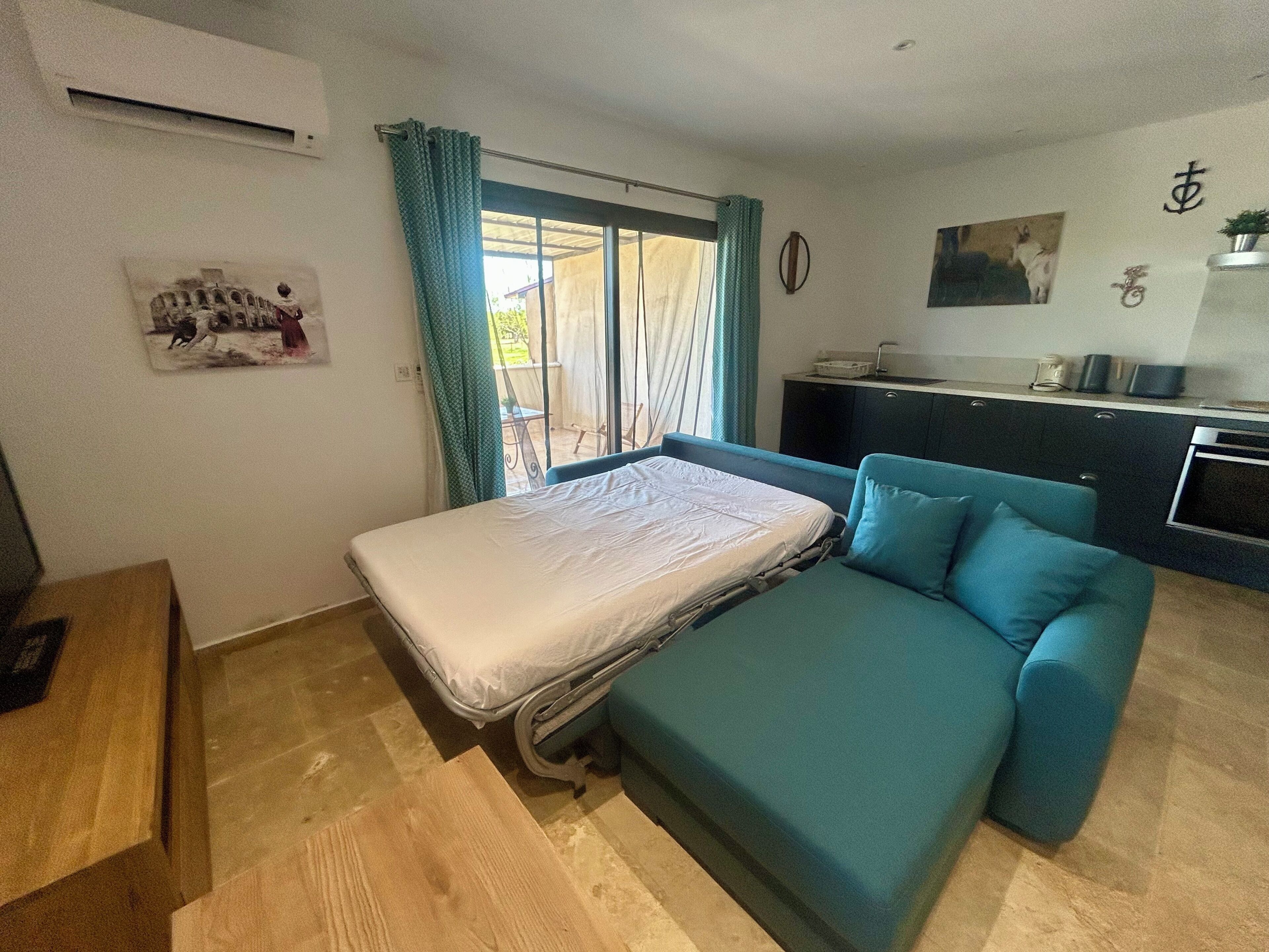 A bedroom with a bed, a sofa, a small desk, a kitchenette, and a sliding glass door to the outside.