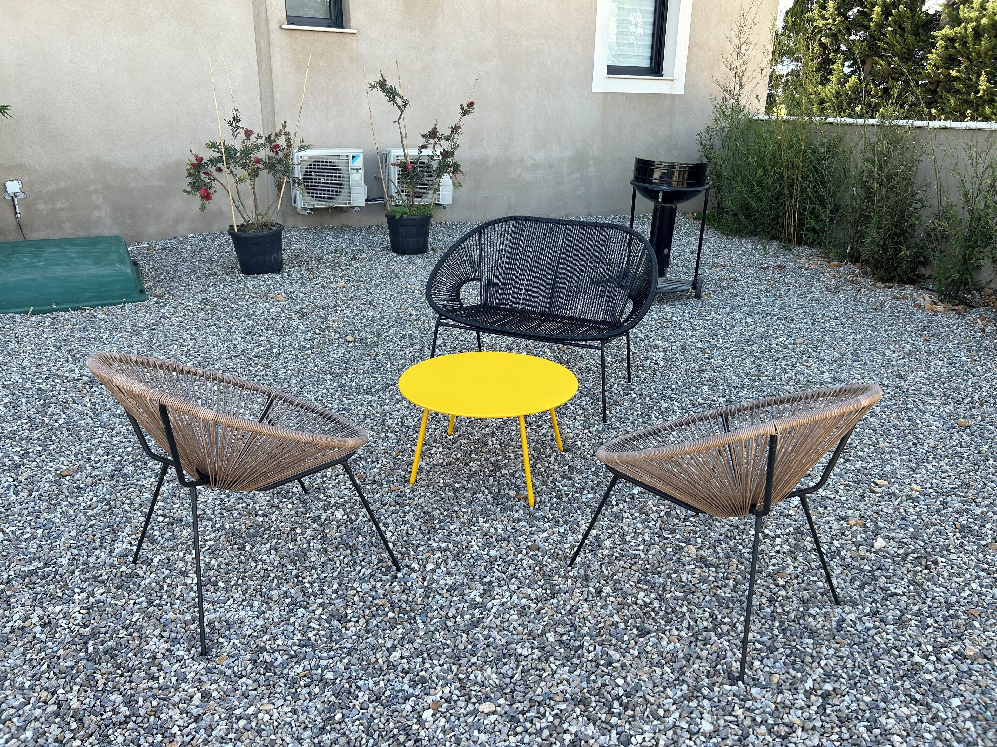 An outdoor seating area with wicker chairs and a yellow table, surrounded by gravel and potted plants.