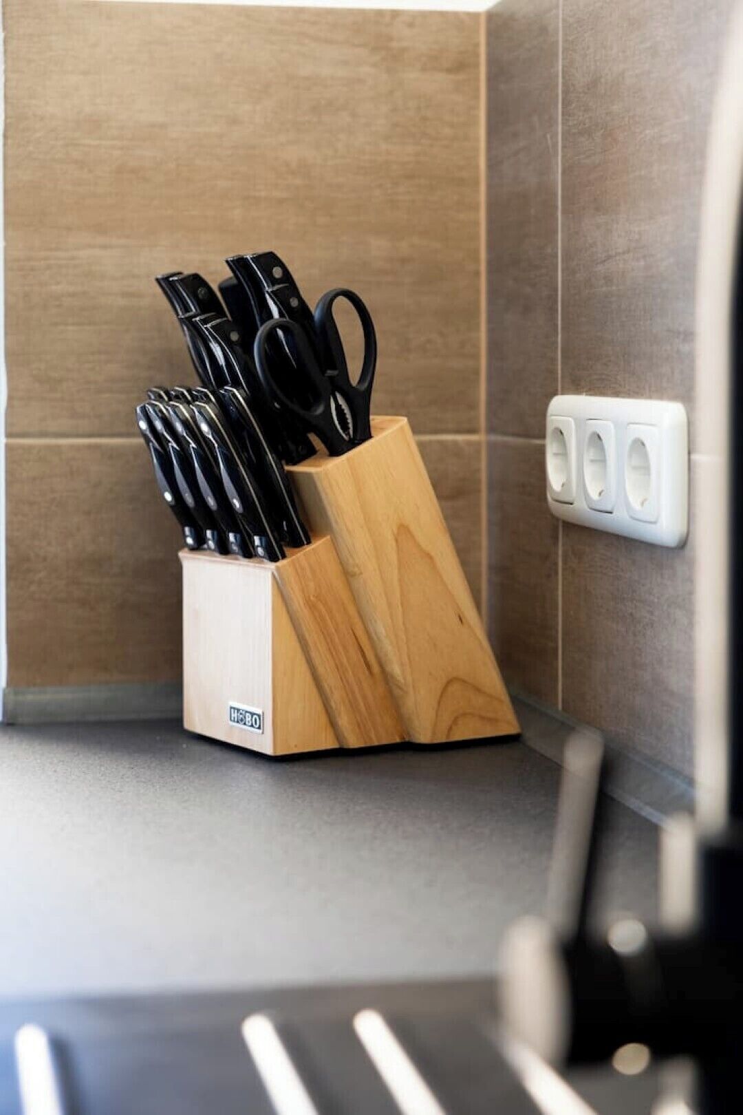 A kitchen corner with a wooden knife block holding various knives and scissors, a power outlet, and a tiled backsplash.