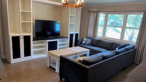 A living room with a sectional sofa, a TV on a stand, white shelving units, and a chandelier.