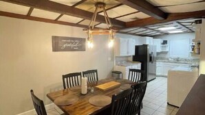A dining area with a wooden table and chairs, a hanging light fixture, and a kitchen in the background.