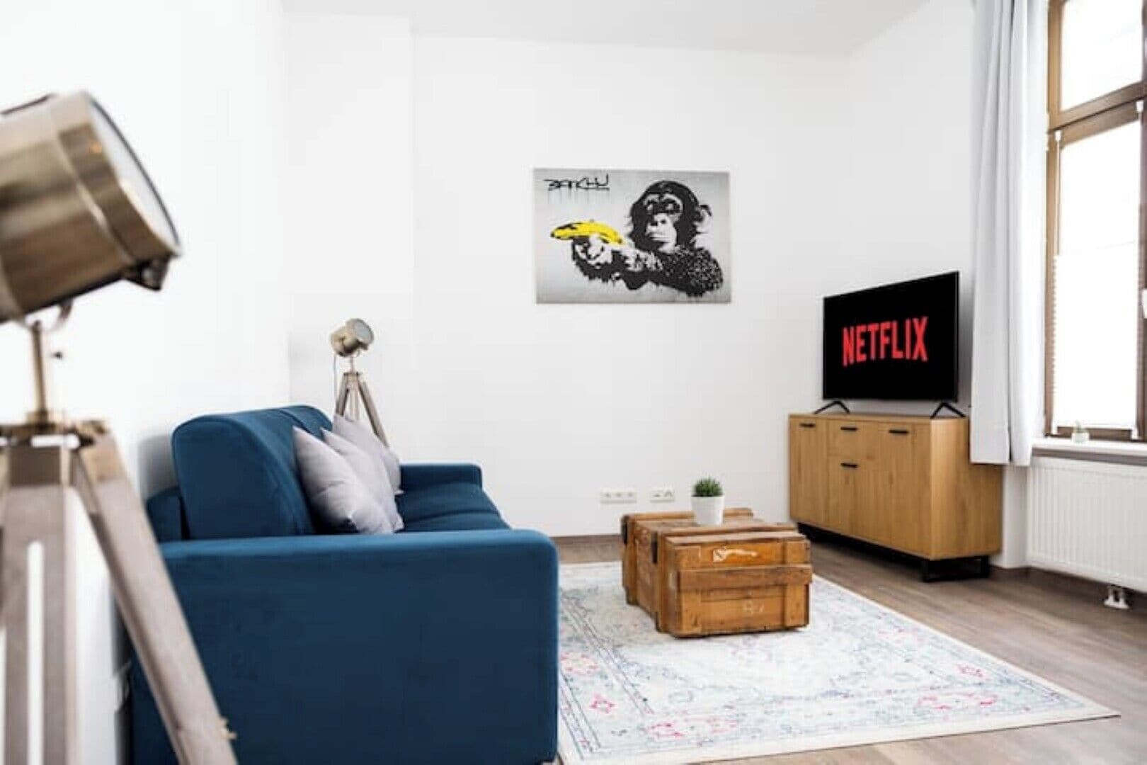 Ein modernes Wohnzimmer mit einem blauen Sofa, einer Holzkommode, einem Fernseher, der 'NETFLIX' zeigt, und einem Wandbild.