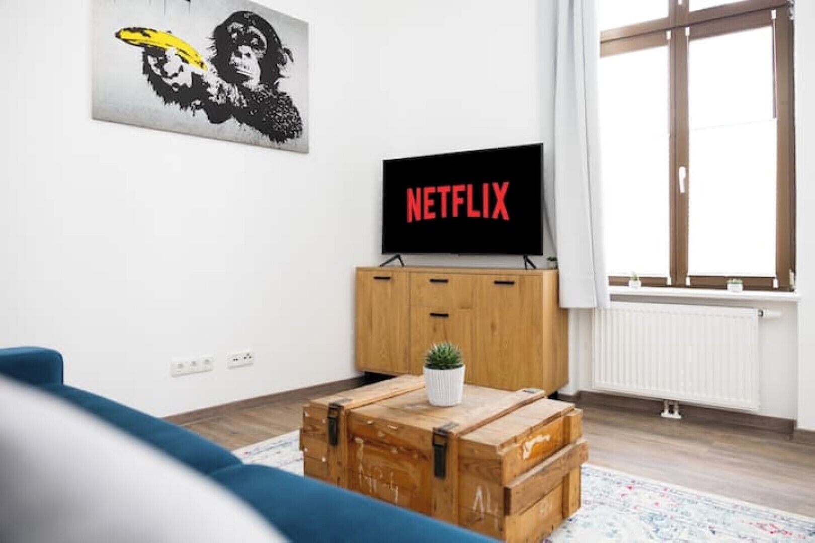 Een woonkamer met een houten kast, een blauwe bank, een houten kist en een tv waarop 'NETFLIX' staat.