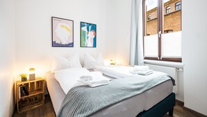 Una cama bien hecha con sábanas blancas, una colcha a rayas y una almohada. Hay dos cuadros en la pared, una mesita de noche con una lámpara y una ventana con persianas.