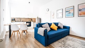 Un salón moderno con un sofá azul, dos sillas blancas, una cocina al fondo y un reloj en la pared.