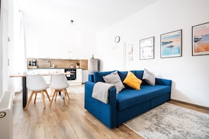 Ein modernes Wohnzimmer mit einem blauen Sofa, zwei weißen Sesseln, einer Küche im Hintergrund und einer Uhr an der Wand.