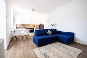 Uma sala de estar moderna com um sofá azul, uma mesa de jantar com cadeiras, uma cozinha ao fundo e um tapete grande no chão.