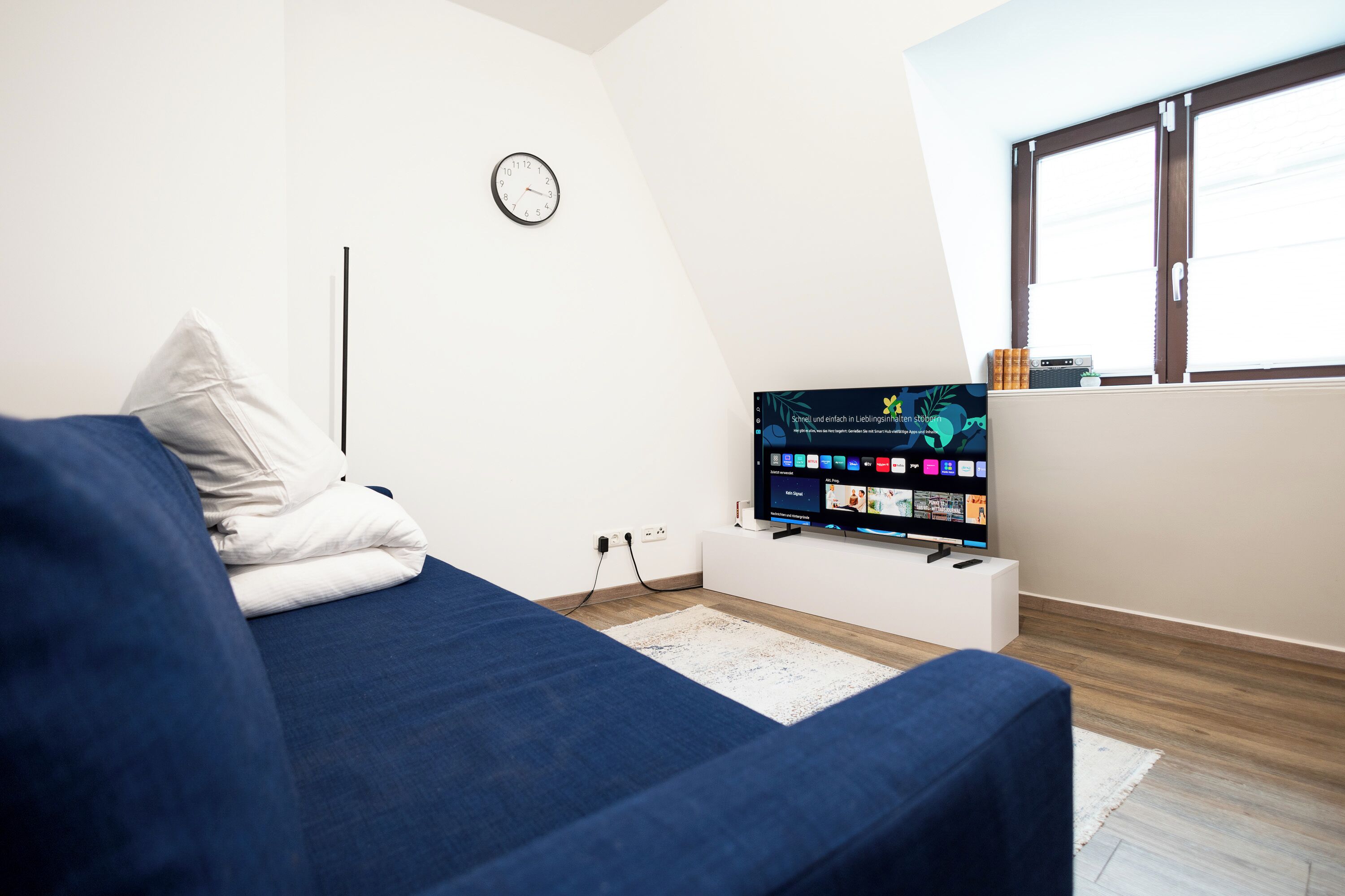 Ein modernes Schlafzimmer mit einem blauen Sofa, einem Flachbildfernseher auf einem weißen Schrank, einer Wanduhr und einem Fenster mit Jalousien.