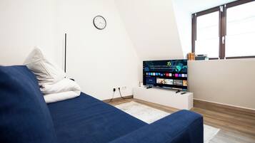 Ein modernes Schlafzimmer mit einem blauen Sofa, einem Flachbildfernseher auf einem weißen Schrank, einer Wanduhr und einem Fenster mit Jalousien.