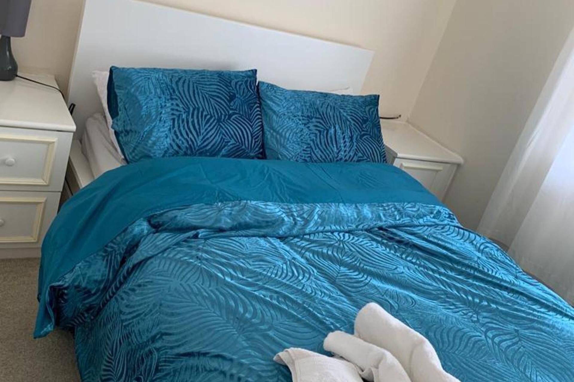A bed with a teal patterned bedspread and matching pillows, a white headboard, and a bedside table with a lamp.