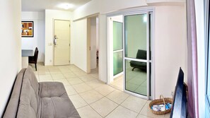 Uma sala de estar com sofá, uma mesa de jantar com cadeiras, uma porta de vidro deslizante para uma varanda e uma televisão.